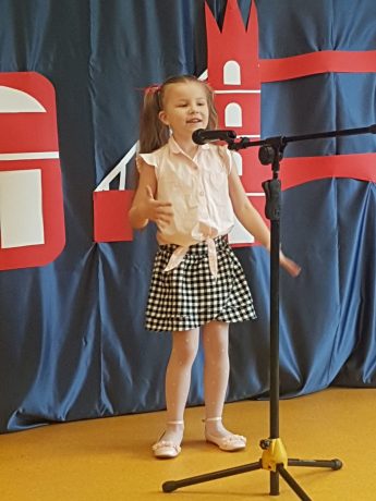Dziewczynka na scenie śpiewa piosenkę. Za nią kotara z elementami przedstawiającymi brytyjskie symbole: piętrowy czerwony autobus, golden bridge i flaga Wielkiej Brytanii. 