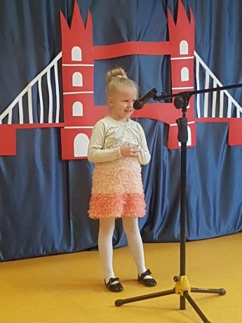 Dziewczynka na scenie śpiewa piosenkę. Za nią kotara z elementami przedstawiającymi brytyjskie symbole: piętrowy czerwony autobus, golden bridge i flaga Wielkiej Brytanii. 