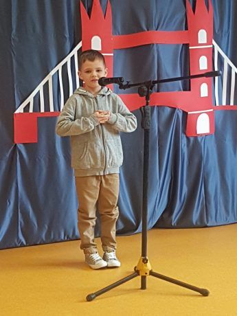 Chłopiec na scenie śpiewa piosenkę. Za nim kotara z elementami przedstawiającymi brytyjskie symbole: piętrowy czerwony autobus, golden bridge i flaga Wielkiej Brytanii. 