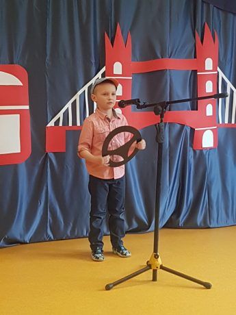 Chłopiec na scenie śpiewa piosenkę. Za nim kotara z elementami przedstawiającymi brytyjskie symbole: piętrowy czerwony autobus, golden bridge i flaga Wielkiej Brytanii. 