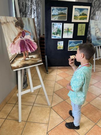 Chłopiec jedząc tort podziwia obraz przedstawiający dziewczynkę w różowej sukience wspinającą się na palce w pozie baletnicy.