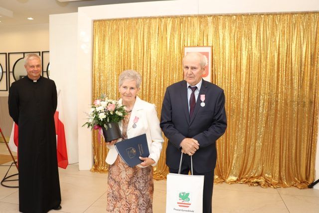 Para odznaczona medalem, trzymająca w ręku kwiaty, list gratulacyjny oraz upominek. Obok stoi ks. Leszek Dąbrowski