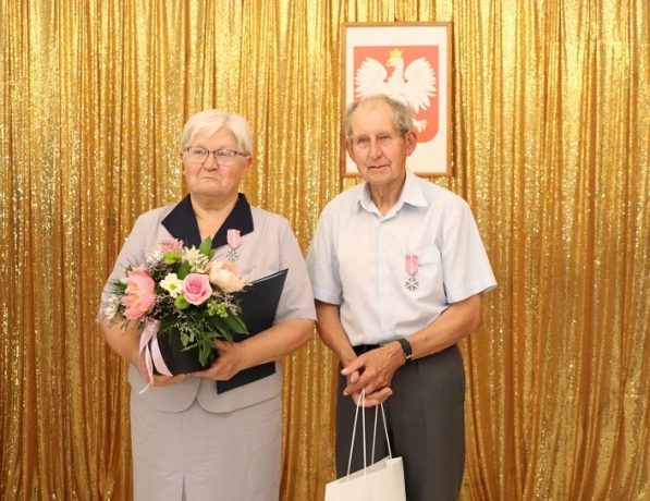Para odznaczona medalem, trzymająca w ręku kwiaty, list gratulacyjny oraz upominek